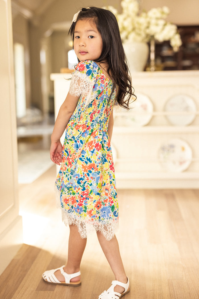 A young girl with long dark hair stands indoors on a wooden floor, glancing over her shoulder at the camera. She wears Ivy City Co's Mini Ada Dress in Vibrant Yarrow Print, featuring bright florals and lace details, paired with white sandals.