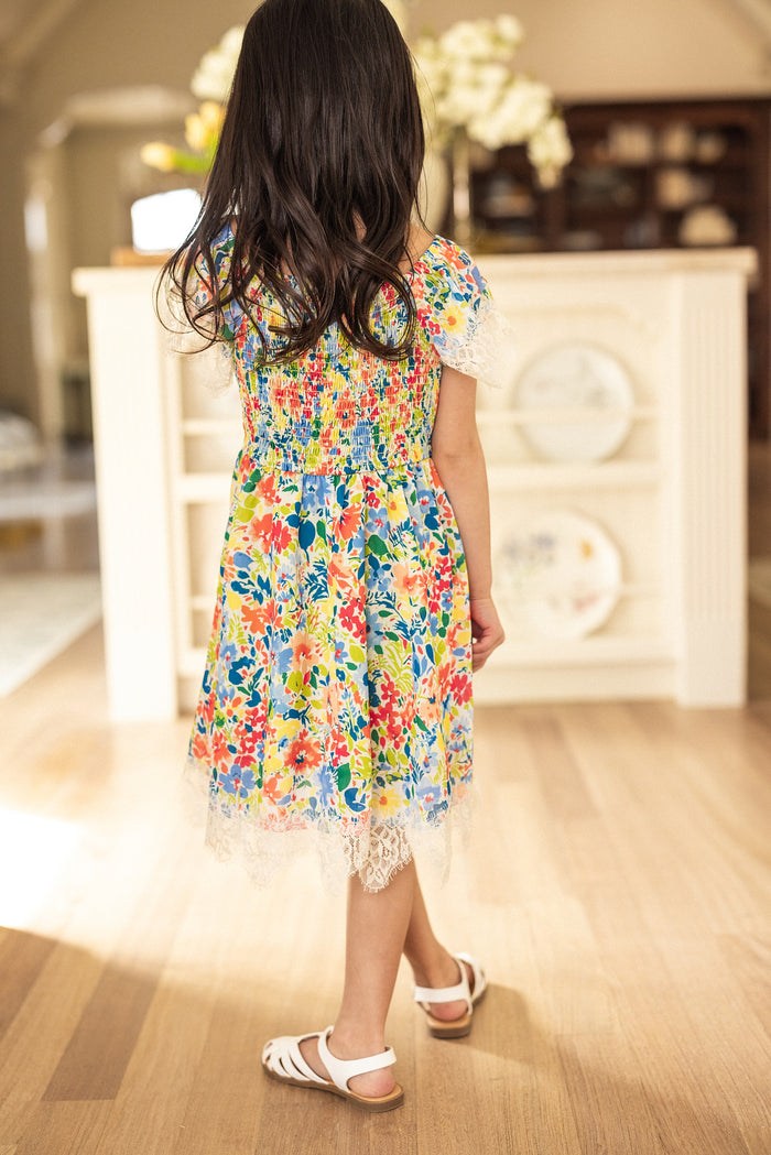 A young girl with long dark hair stands indoors on a wooden floor, facing away from the camera, wearing the Ivy City Co Mini Ada Dress in Vibrant Yarrow Print and white sandals.