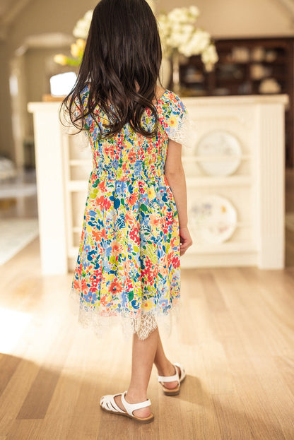 A young girl with long dark hair stands indoors on a wooden floor, facing away from the camera, wearing the Ivy City Co Mini Ada Dress in Vibrant Yarrow Print and white sandals.