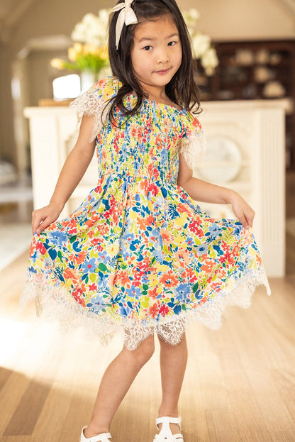 A young girl poses indoors wearing the Ivy City Co Mini Ada Dress in Vibrant Yarrow Print, featuring a colorful floral chiffon design with delicate lace trim. She accessorizes with a white bow in her hair and white shoes, standing on a wooden floor against a softly blurred background.