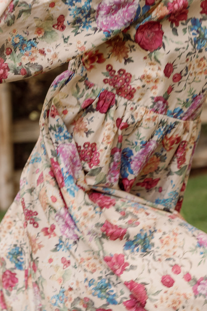 Close-up of the Ivy City Co Mini Adelpha Dress in Autumn Amity Floral, highlighting its flowing fabric as it moves outdoors. The dress boasts a lively floral pattern in shades of pink, red, blue, and green against a beige backdrop. Textured details such as a smocked bodice and delicate balloon sleeves enhance the vibrant appeal of this style.