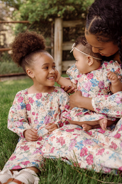 A woman holds a baby and sits beside a young girl, all three smiling at each other while sitting on green grass outdoors. They are wearing matching Ivy City Co Mini Adelpha Dresses in Autumn Amity Floral, featuring playful balloon sleeves and smocked bodice details.