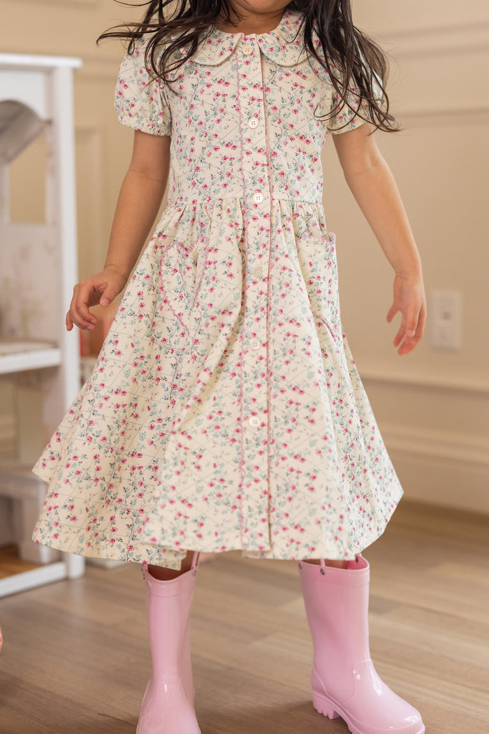 A young child in the Ivy City Co Mini Alayna Play Dress in Ivory Pansy Trellis and pink rain boots stands on a wooden floor indoors, with her face not visible.