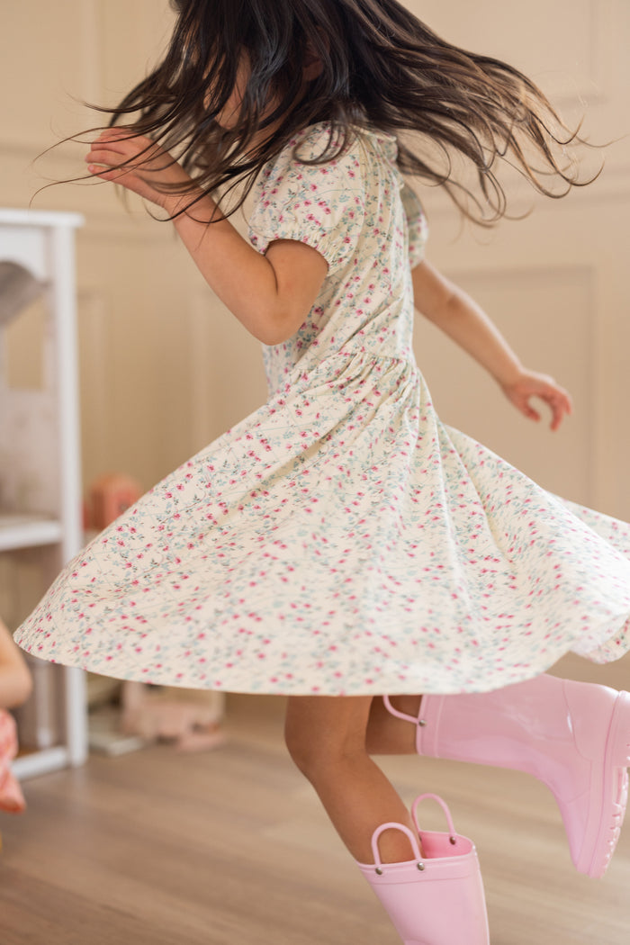 A young girl twirls indoors in the Ivy City Co Mini Alayna Play Dress in Ivory Pansy Trellis, her long hair flying and pink rain boots peeking out as she spins across the wooden floor in comfy cotton-spandex knit.