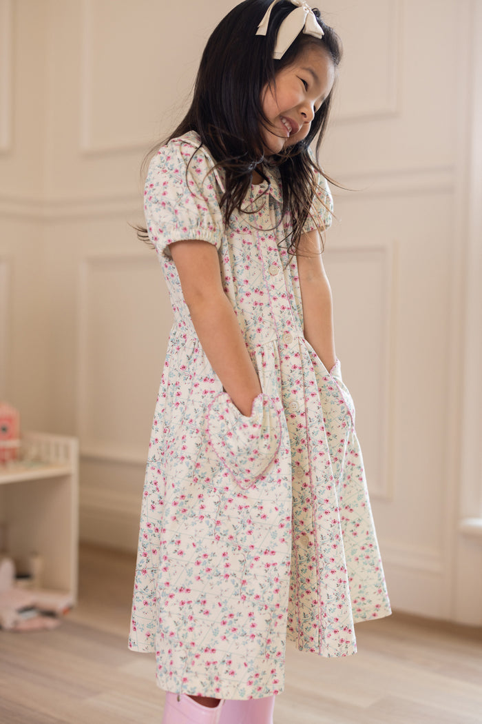 A young girl with long dark hair wears the Ivy City Co Mini Alayna Play Dress in Ivory Pansy Trellis, featuring puff sleeves and a bow headband. The soft cotton-spandex dress has pockets and is shown indoors against light-colored walls.