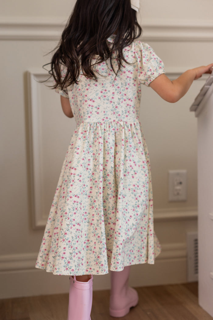 A young girl with long dark hair, wearing the Ivy City Co Mini Alayna Play Dress in Ivory Pansy Trellis and pink rain boots, stands facing away near a white wall and a piece of furniture.