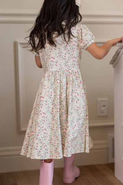 A young girl with long dark hair, wearing the Ivy City Co Mini Alayna Play Dress in Ivory Pansy Trellis and pink rain boots, stands facing away near a white wall and a piece of furniture.