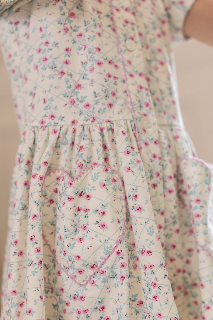 Close-up of Ivy City Co's Mini Alayna Play Dress in Ivory Pansy Trellis, a light cotton-spandex spring dress with pink and blue floral print, two heart-shaped pockets outlined in pink, and softly gathered waist.