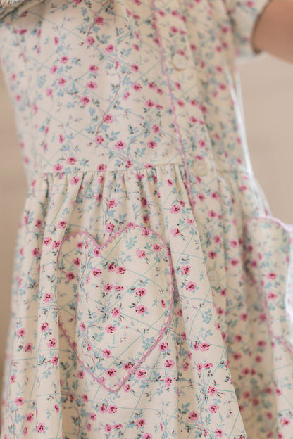 Close-up of Ivy City Co's Mini Alayna Play Dress in Ivory Pansy Trellis, a light cotton-spandex spring dress with pink and blue floral print, two heart-shaped pockets outlined in pink, and softly gathered waist.