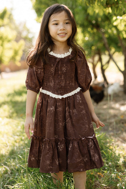 A young girl with long dark hair is wearing the Ivy City Co Mini Asha Dress in Warm Cocoa Valencia Toile, which features balloon sleeves and a smocked bodice, as she stands smiling outdoors on grass with green trees and sunlight in the background.