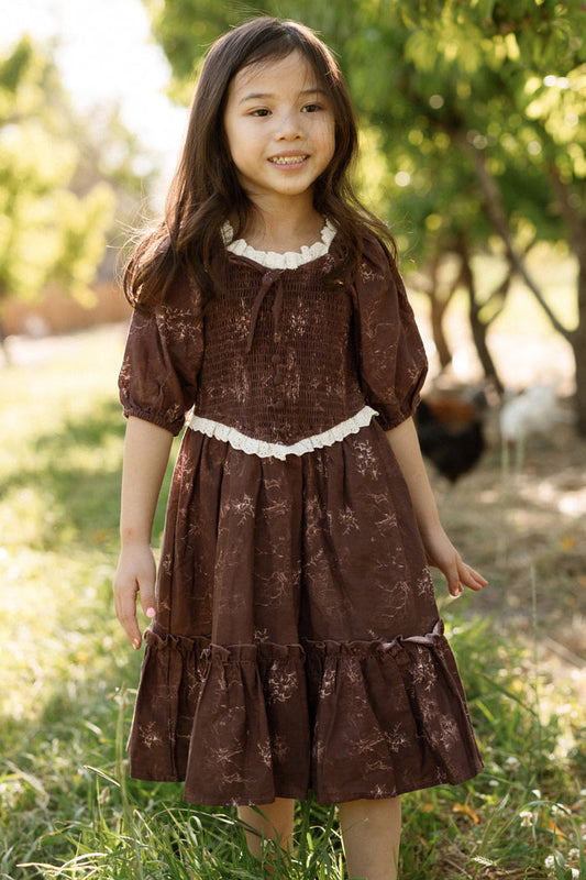 A young girl with long dark hair is wearing the Ivy City Co Mini Asha Dress in Warm Cocoa Valencia Toile, which features balloon sleeves and a smocked bodice, as she stands smiling outdoors on grass with green trees and sunlight in the background.