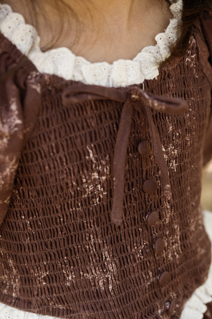 Close-up of a child wearing the Ivy City Co Mini Asha Dress in Warm Cocoa Valencia Toile, featuring a smocked bodice, white lace collar, fabric buttons, and a tied bow at the neckline. Only part of the neck and shoulders are visible.