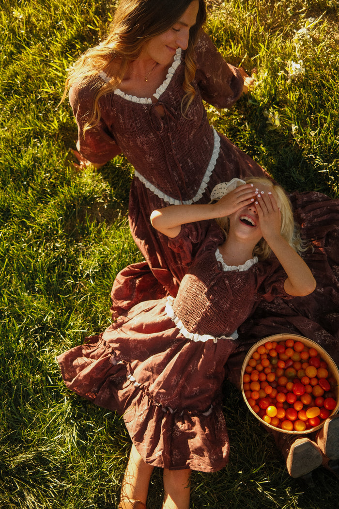 A woman and a laughing child sit on the grass beside a basket of cherry tomatoes, both dressed in matching Ivy City Co Mini Asha Dresses in Warm Cocoa Valencia Toile, featuring smocked bodices and playful balloon sleeves. The child covers their eyes as sunlight brightens the scene.
