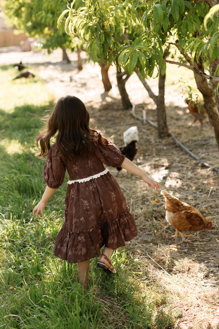 A young girl in the Ivy City Co Mini Asha Dress in Warm Cocoa Valencia Toile, featuring balloon sleeves and a smocked bodice, walks barefoot on grass near chickens beneath leafy trees on a sunny day.