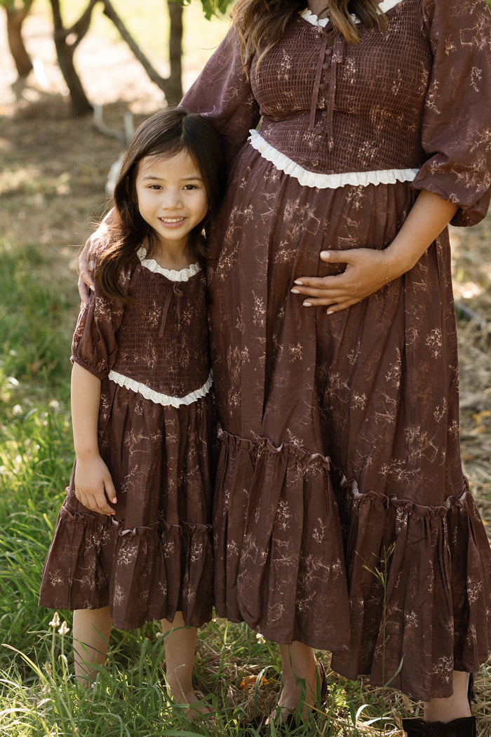 A woman and a young girl stand outdoors on green grass among trees, both wearing matching Ivy City Co Mini Asha Dresses in Warm Cocoa Valencia Toile with white trim. The woman cradles her pregnant belly, highlighting the dress’s smocked bodice and elegant balloon sleeves, while the girl smiles beside her.