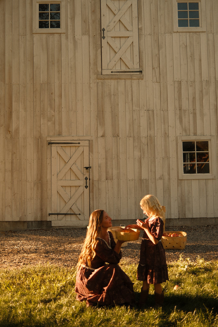 A woman kneels on the grass, handing a wrapped gift to a young girl outside a wooden barn in warm sunlight. Both appear joyful as they share a moment together, dressed in matching Ivy City Co Mini Asha Dresses in Warm Cocoa Valencia Toile with balloon sleeves and smocked bodices.