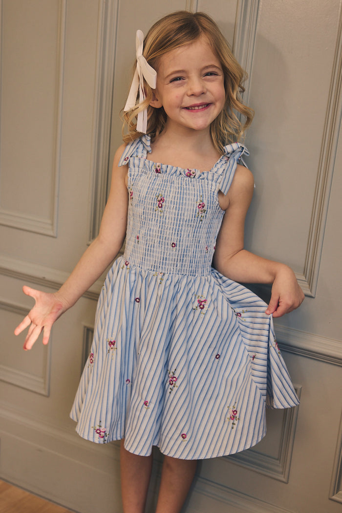A young girl with blonde hair and a white bow smiles in the Ivy City Co Mini Bluebell Dress in Blue & White Stripes, featuring a charming smocked bodice, as she poses against a paneled wall.