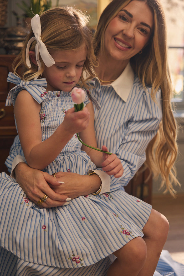 A woman and a young girl sit together, both wearing Ivy City Co's Mini Bluebell Dress in Blue & White Stripes. The girl has a white ribbon in her hair and gazes at a pink flower as sunlight softly fills the room.