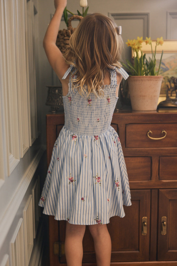 A young girl in Ivy City Co's Mini Bluebell Dress in Blue & White Stripes stands facing a wooden sideboard, reaching toward a tall basket, with potted yellow daffodils displayed on the sideboard.
