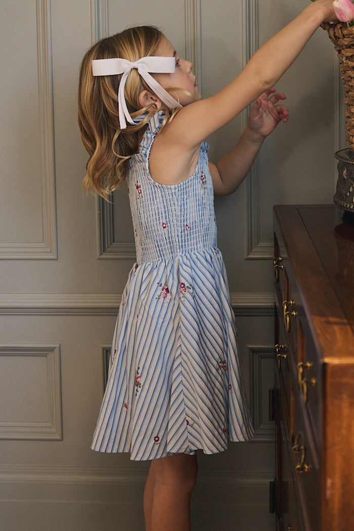 A young girl with shoulder-length hair and a pink ribbon reaches for a woven basket on a wooden dresser, wearing the Ivy City Co Mini Bluebell Dress in Blue & White Stripes, adorned with delicate small floral patterns.