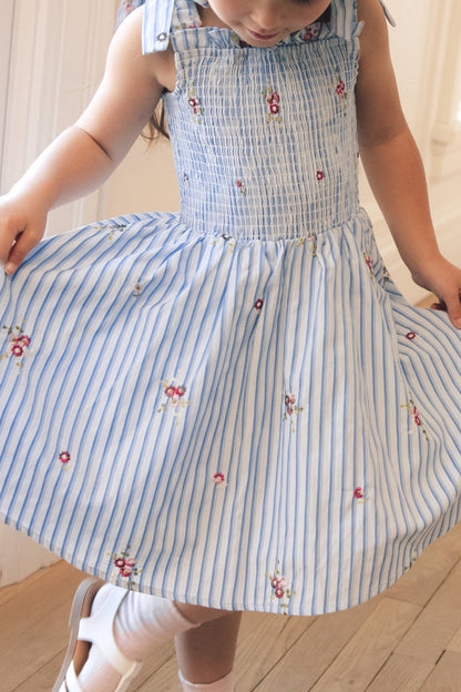 A young child twirls in the Ivy City Co Mini Bluebell Dress in Blue & White Stripes, featuring small florals, a smocked bodice, and tied shoulder straps, paired with white shoes and socks on a wooden floor.