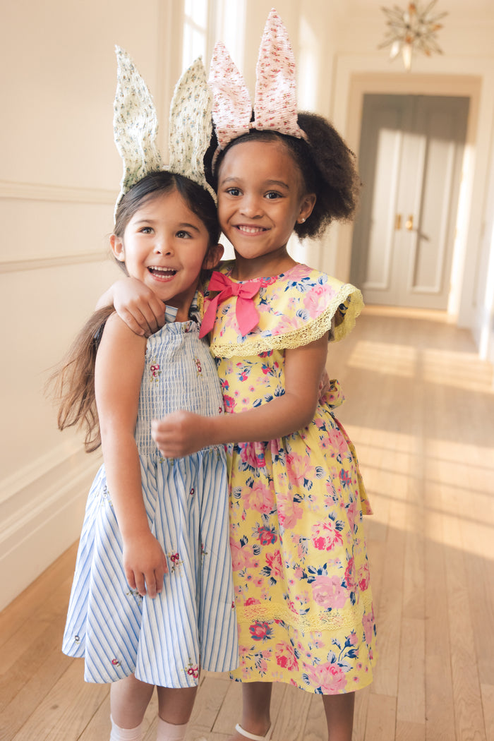 Two young girls wear Ivy City Co’s Mini Bluebell Dress in Blue & White Stripes, smiling and hugging each other in a sunlit hallway, their outfits brightening the cheerful scene.