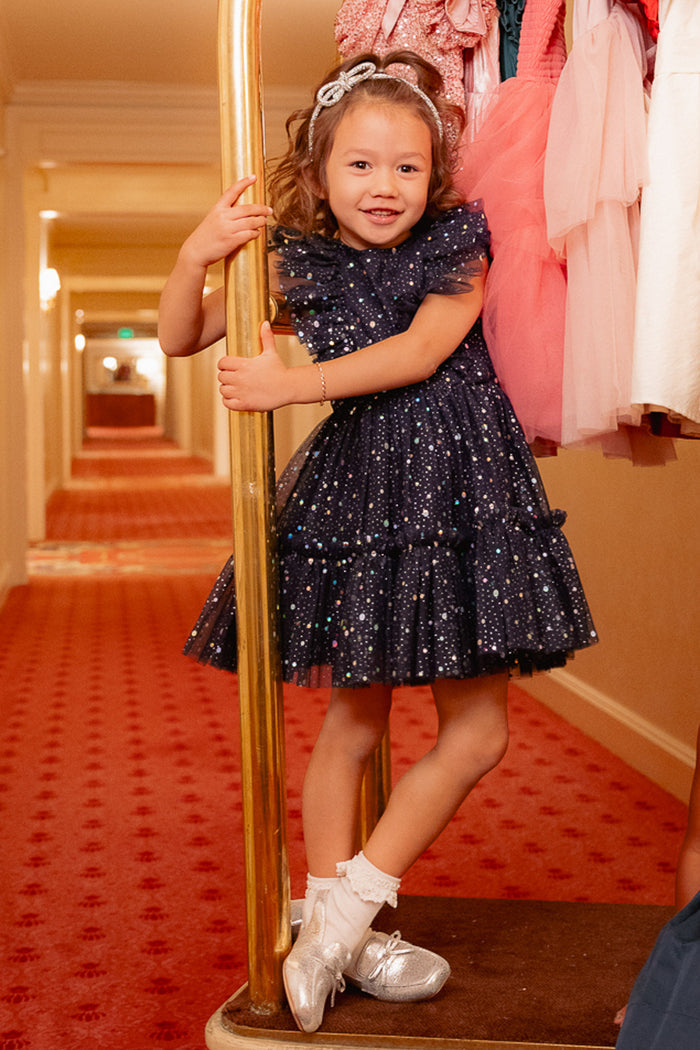 A young girl in the Ivy City Co Mini Brynlee Dress in Sparkling Stars Navy and silver shoes smiles on a hotel luggage cart, holding a gold pole, with pink dresses hanging beside her in the carpeted hallway.