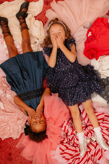 Two young girls in fancy dresses, including the Ivy City Co Mini Brynlee Dress in Sparkling Stars Navy, smile and play on a colorful pile of fabrics atop a red carpet, surrounded by vibrant textures.