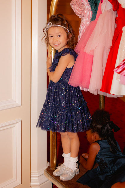 A young girl in the Ivy City Co Mini Brynlee Dress in Sparkling Stars Navy and silver shoes smiles while standing on a hotel luggage cart. A second child in a dark winter dress sits below, with colorful dresses hanging above them.