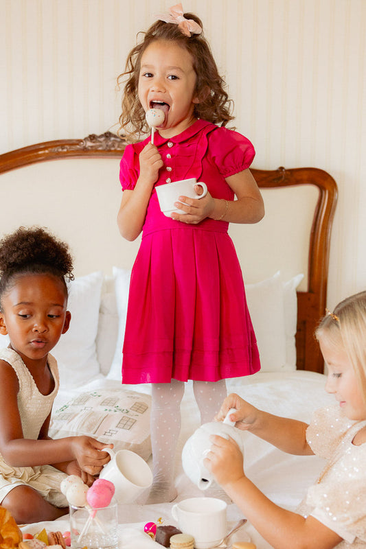 Three young girls have a tea party on a bed. The center girl stands and smiles, wearing the Ivy City Co Mini Cassian Dress in Hot Pink, while holding a teacup and treat. The other two pour tea and play with desserts.