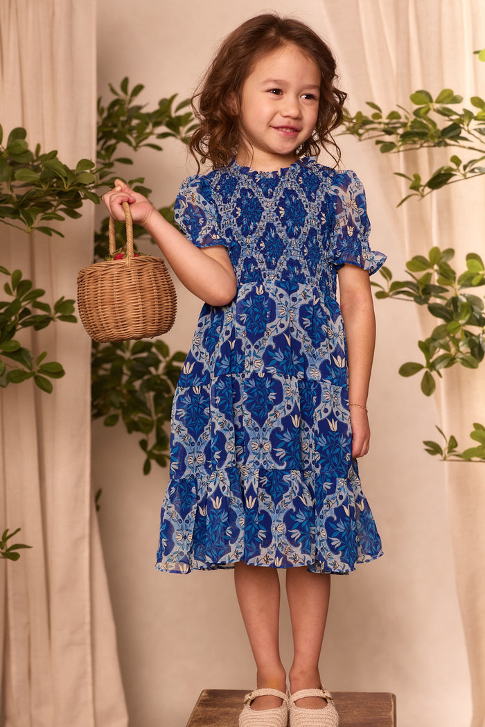 A young girl wears the Ivy City Co Mini Cordelia Dress in Trellis Foulard, standing on a wooden box and holding a wicker basket. Green leafy plants and cream curtains create a charming backdrop.