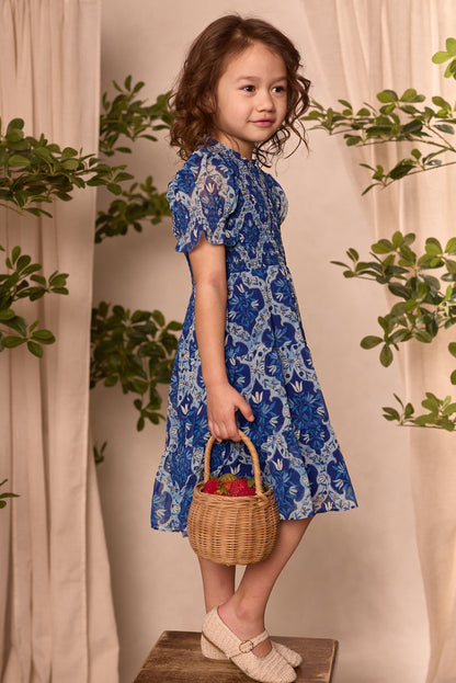 A young girl with wavy brown hair stands on a wooden box, wearing the Ivy City Co Mini Cordelia Dress in Trellis Foulard and cream shoes. She holds a wicker basket with red flowers. Beige curtains and green plants are behind her.