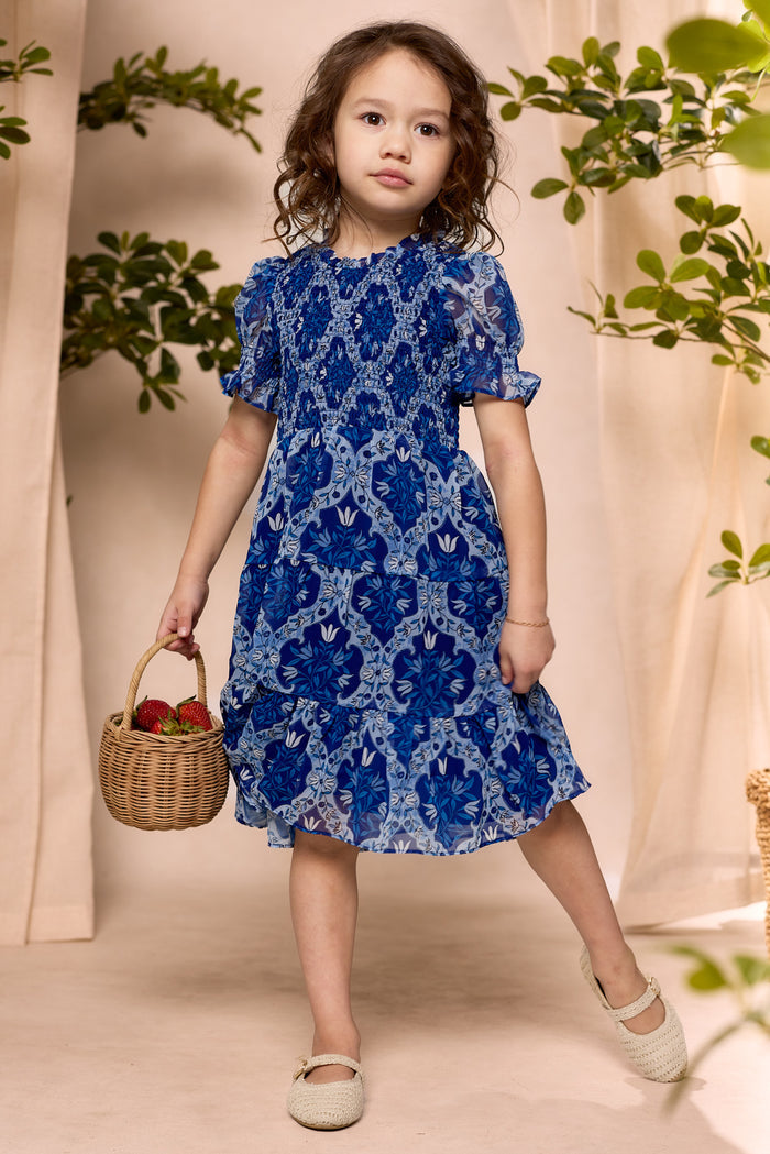 A young girl wears the Ivy City Co Mini Cordelia Dress in Trellis Foulard with beige shoes, holding a wicker basket of strawberries indoors. Green plants and neutral curtains are in the background.