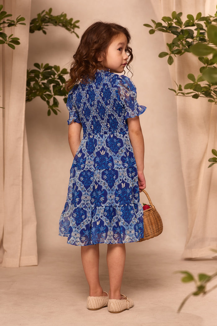 A young girl with curly brown hair wears the Ivy City Co Mini Cordelia Dress in Trellis Foulard and beige shoes, holding a small woven basket. She looks over her shoulder with green plants and beige curtains in the background.