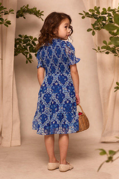 A young girl with curly brown hair wears the Ivy City Co Mini Cordelia Dress in Trellis Foulard and beige shoes, holding a small woven basket. She looks over her shoulder with green plants and beige curtains in the background.
