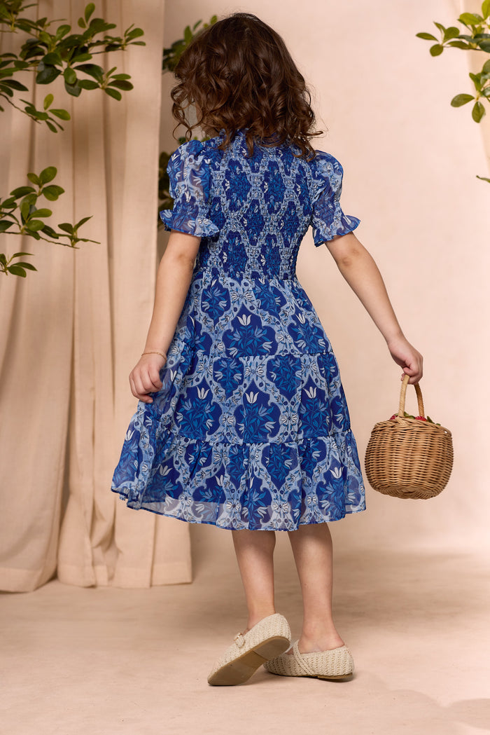 A young girl with curly hair, seen from behind in the Ivy City Co Mini Cordelia Dress in Trellis Foulard, carries a small wicker basket on a light floor with green leaves and beige curtains—perfect for matching family outfits.