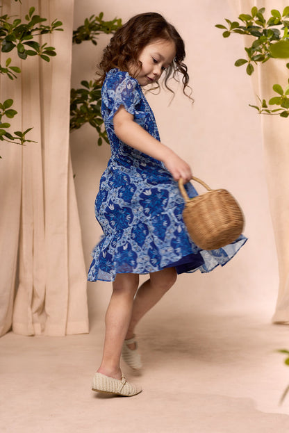 A young girl with brown curly hair twirls joyfully in the Ivy City Co Mini Cordelia Dress in Trellis Foulard, barefoot and holding a woven basket, surrounded by green plants against a light, neutral background.