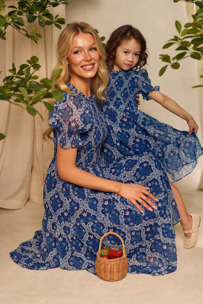 A woman and a young girl in matching Ivy City Co outfits pose indoors; the girl wears the Mini Cordelia Dress in Trellis Foulard as they kneel by strawberries, framed by green branches, showing off their stylish blue floral look.