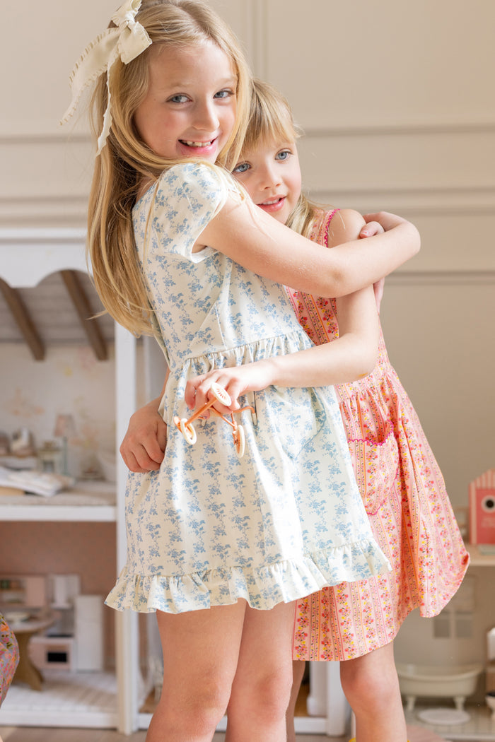 Two young girls smiling and hugging indoors. One is wearing the Ivy City Co Mini Dream Play Dress in Ivory Porcelain Fleur, and the other wears a pink play dress. Both look happy and playful in a cozy room.