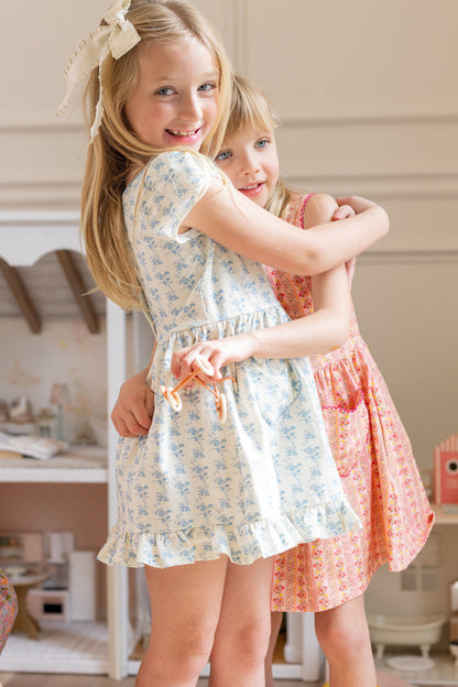 Two young girls smiling and hugging indoors. One is wearing the Ivy City Co Mini Dream Play Dress in Ivory Porcelain Fleur, and the other wears a pink play dress. Both look happy and playful in a cozy room.