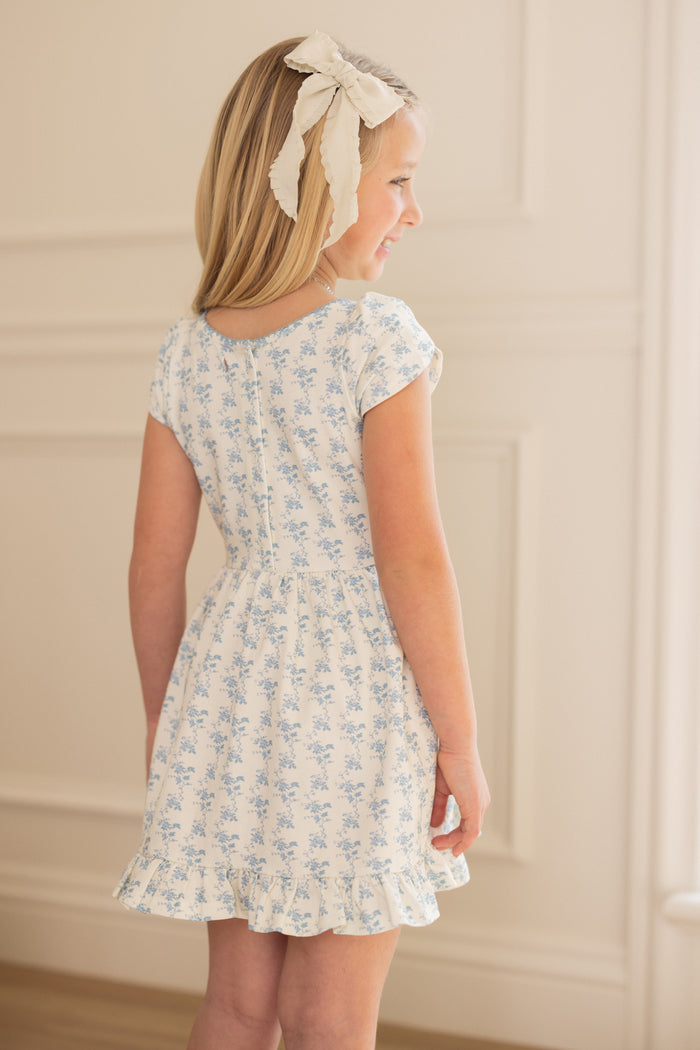 A blonde girl wears the Ivy City Co Mini Dream Play Dress in Ivory Porcelain Fleur and a large white hair bow, standing indoors and smiling slightly away, with light walls and decorative molding in the background.