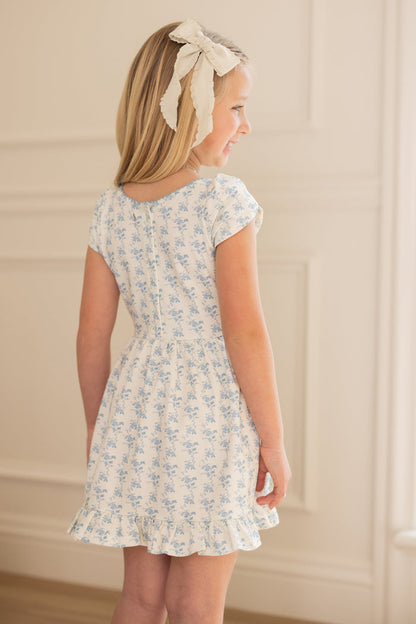 A blonde girl wears the Ivy City Co Mini Dream Play Dress in Ivory Porcelain Fleur and a large white hair bow, standing indoors and smiling slightly away, with light walls and decorative molding in the background.