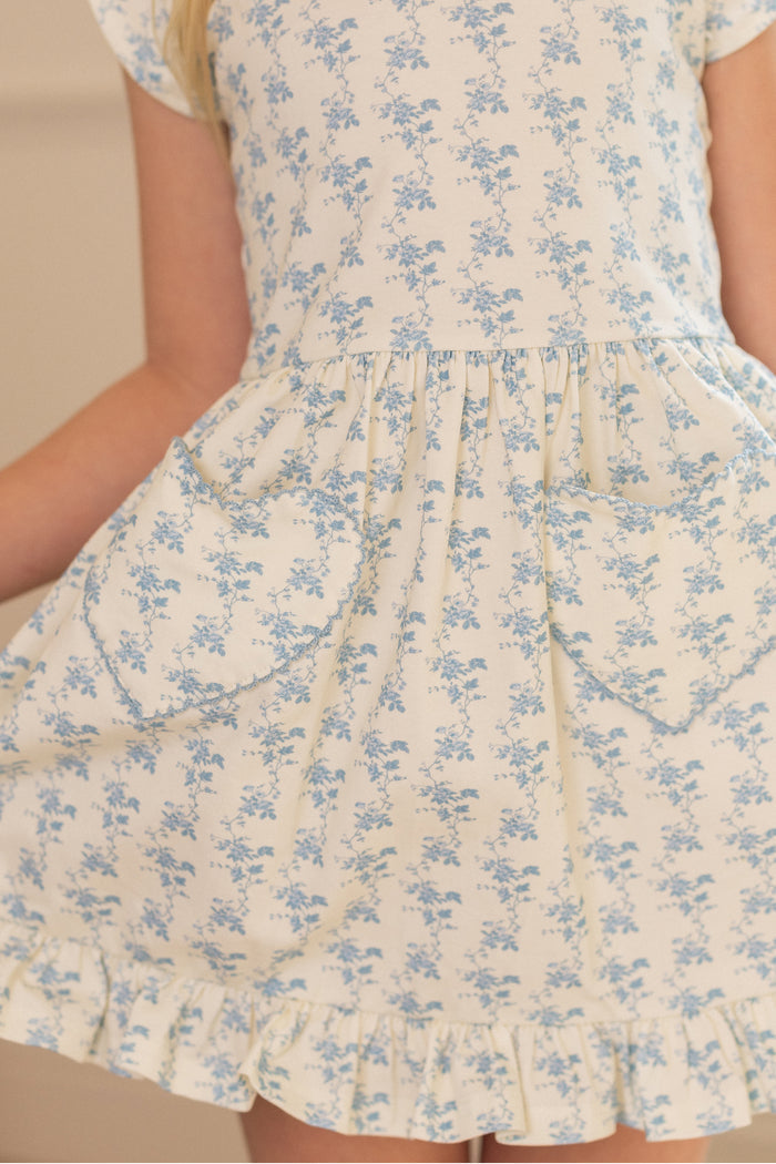 A close-up of a girl wearing the Ivy City Co Mini Dream Play Dress in Ivory Porcelain Fleur, featuring blue floral stretch fabric, ruffled hem, and two heart-shaped front pockets. Only the lower torso and arms are visible.