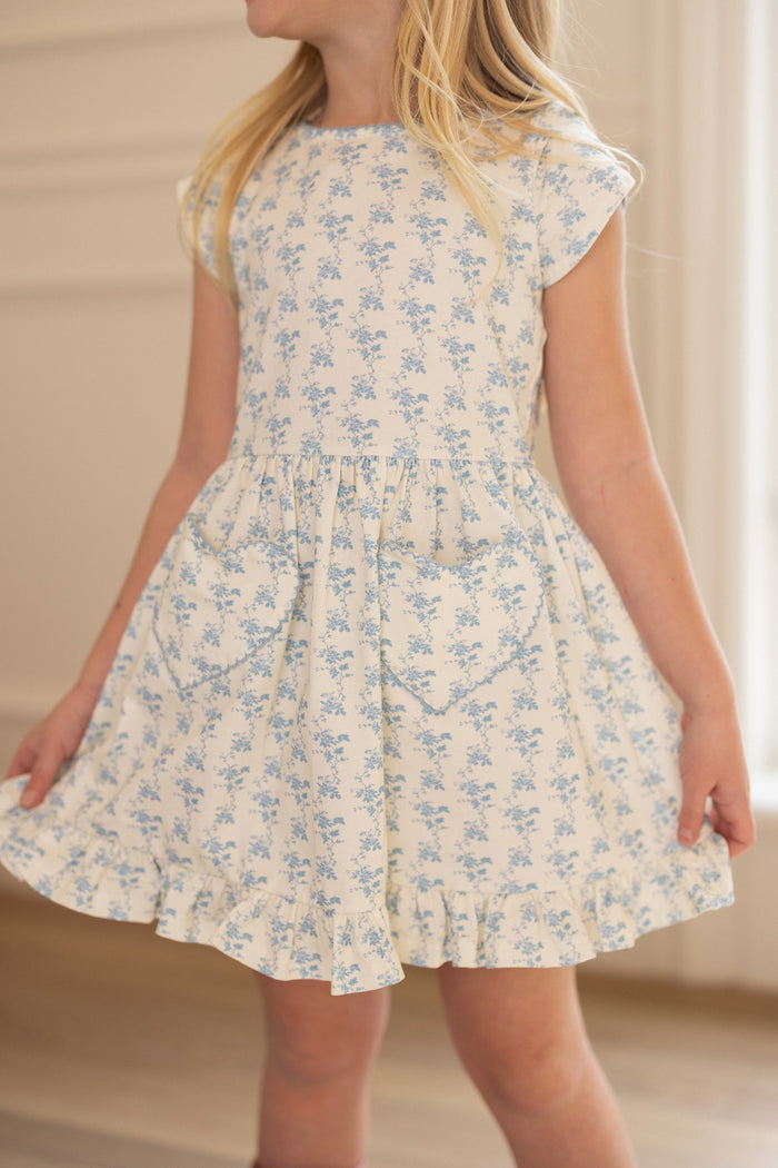 A young girl stands indoors, holding out the sides of her skirt while wearing the Ivy City Co Mini Dream Play Dress in Ivory Porcelain Fleur—a light spring dress for girls featuring blue floral patterns and short sleeves. Her face is turned away from the camera.