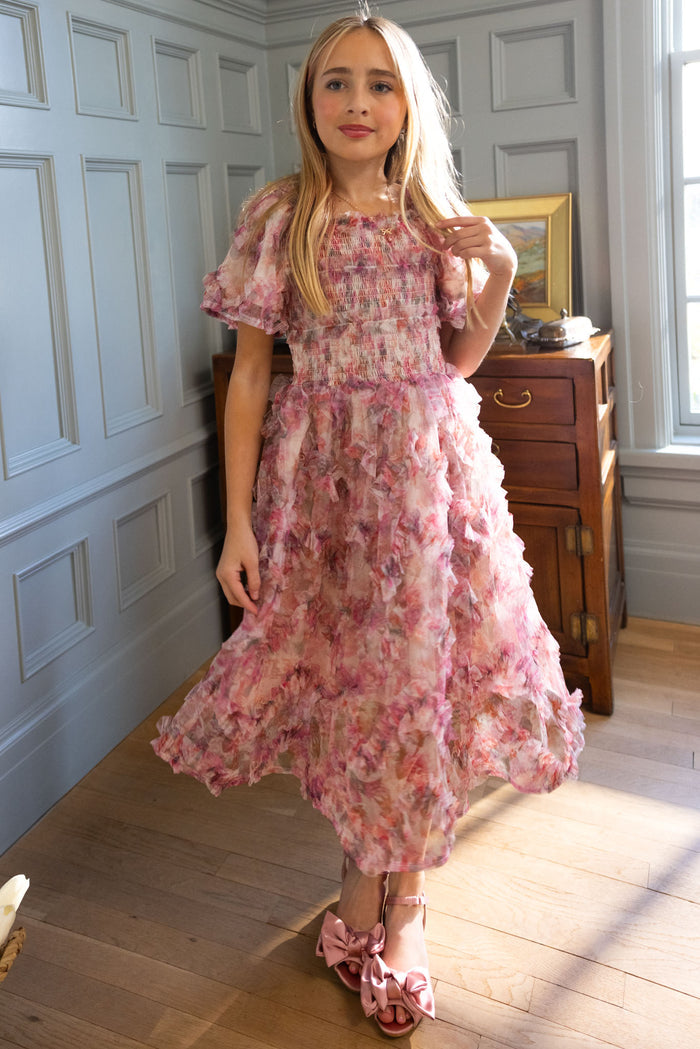 A young girl poses indoors in Ivy City Co's Mini Florentina Dress in Alluring Floral, a pink ruffled style with bow shoes—ideal for spring gatherings. Soft window light highlights her long blonde hair as she stands gracefully.