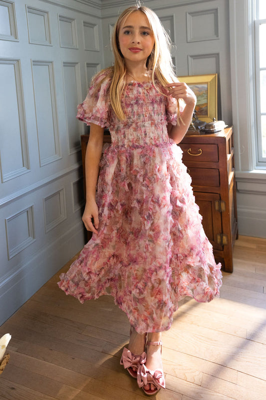 A young girl poses indoors in Ivy City Co's Mini Florentina Dress in Alluring Floral, a pink ruffled style with bow shoes—ideal for spring gatherings. Soft window light highlights her long blonde hair as she stands gracefully.
