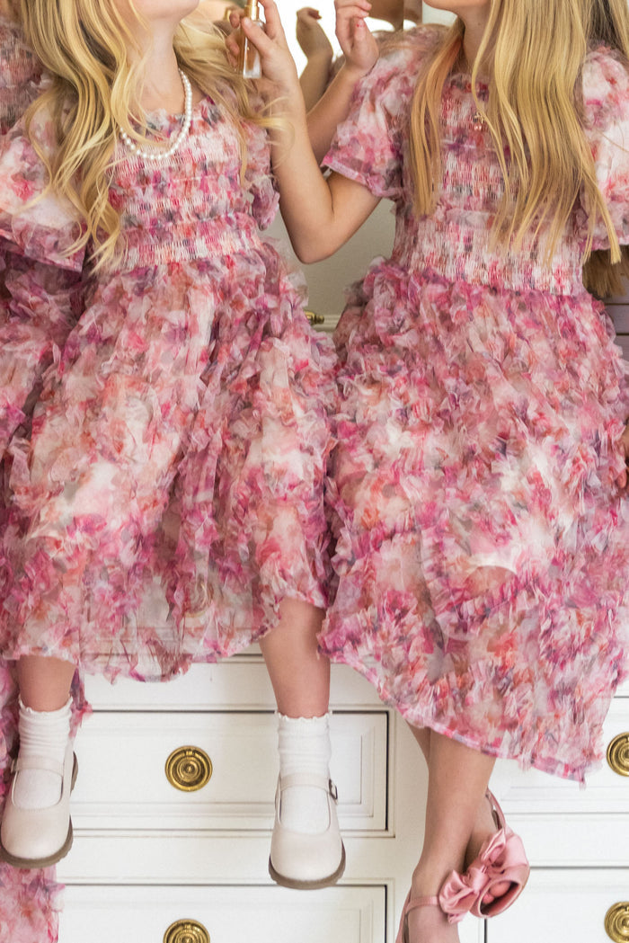 Two young girls with long blonde hair sit on a white dresser, wearing matching Mini Florentina Dresses in Alluring Floral by Ivy City Co. Dressed in white or pink shoes, they’re ready for spring gatherings. The girls’ faces are not visible.
