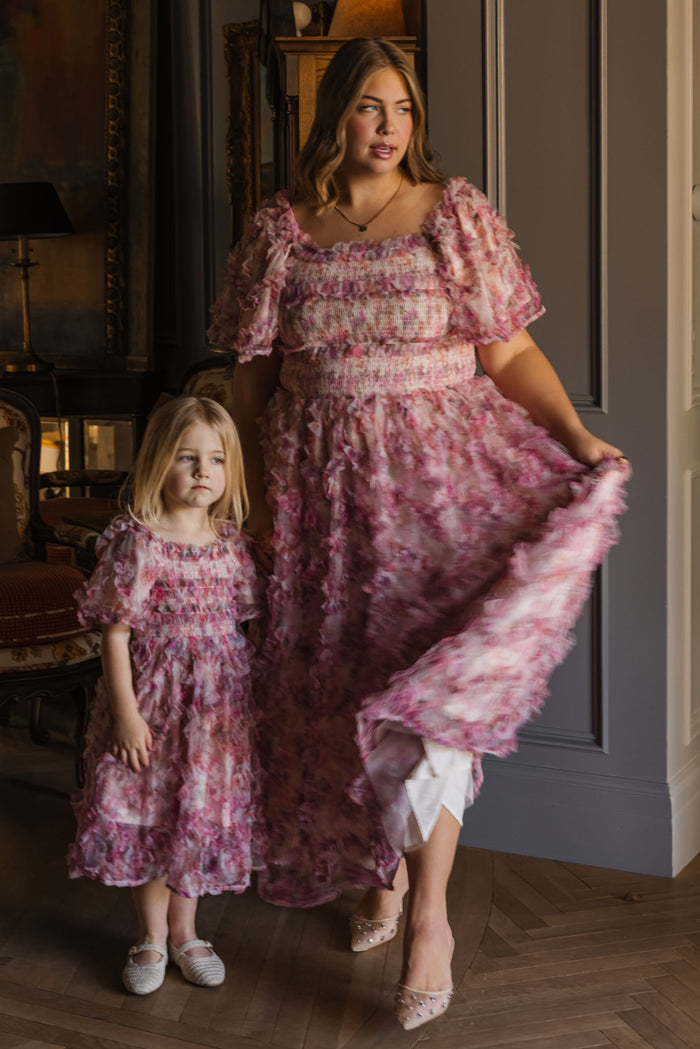 A woman and young girl, both with light brown hair, wear matching Ivy City Co Mini Florentina Dresses in Alluring Floral—ideal for spring gatherings. The woman holds her dress while the girl stands beside her, both barefoot or in light shoes.