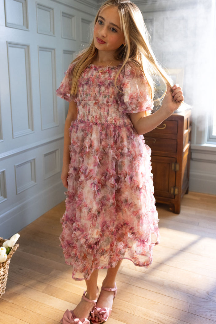 A young girl stands indoors in the Ivy City Co Mini Florentina Dress in Alluring Floral—a pink and purple, ruffle-detailed dress perfect for spring gatherings—gently touching her long blonde hair near a wooden cabinet by a sunlit window.