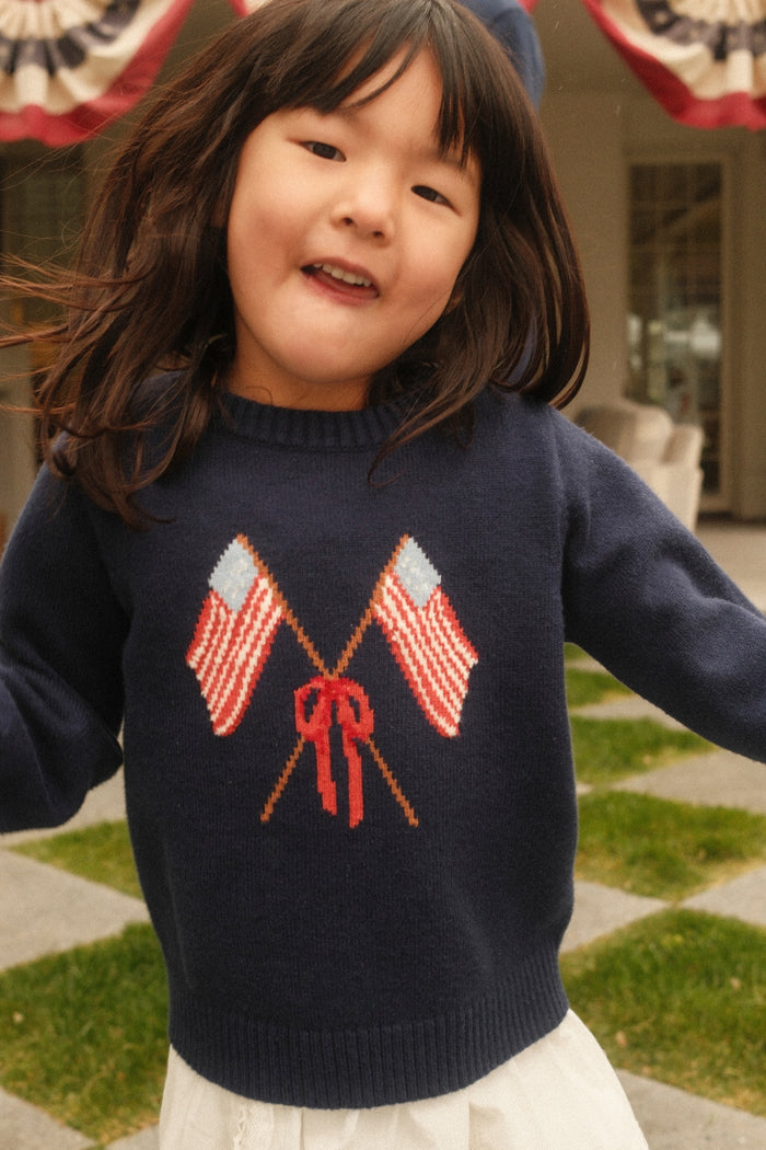 A young girl with long dark hair poses outdoors in Ivy City Co’s Mini Freedom Sweater in Midnight, featuring two crossed American flags and a red bow. She stands on a checkered lawn with patriotic banners, perfect for the Fourth of July.